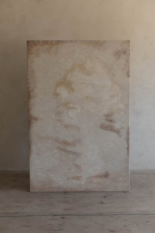 Art By Suzette Newman is a handmade, large rectangular canvas with textured neutral tones—light beige, cream, and brown patches—standing upright against a softly lit wall and cement floor, showcasing premium craftsmanship.