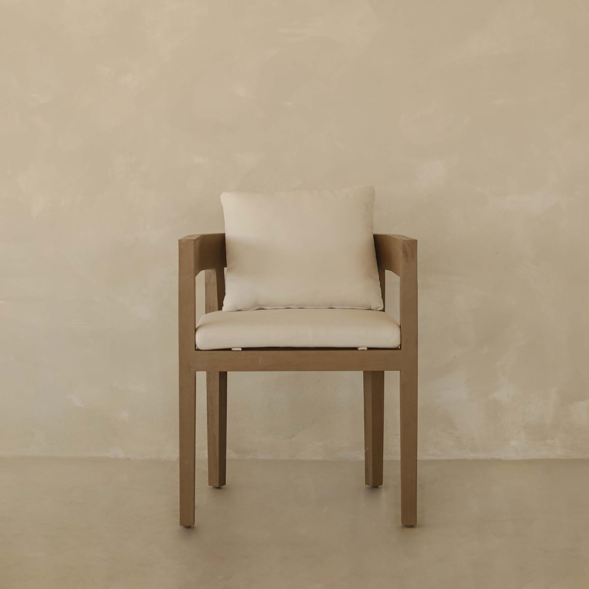 The Astier Outdoor Dining Chair, featuring a minimalist wooden frame with a beige cushion and back pillow, rests against a textured beige wall and floor, showcasing California sophistication and durable artisanal craftsmanship.