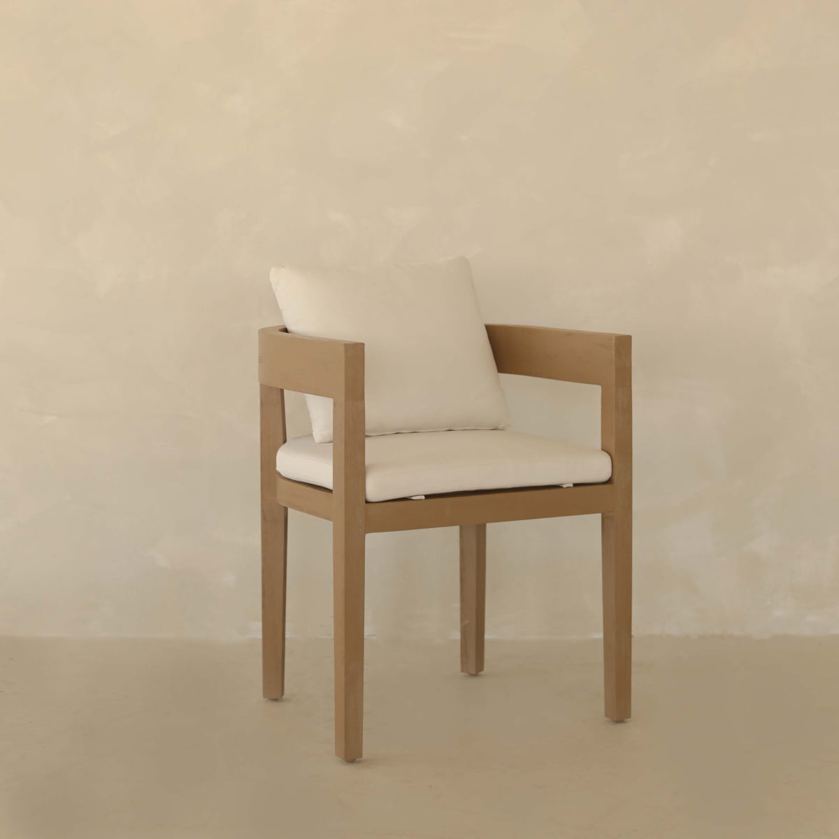The Astier Outdoor Dining Chair, with its light wood frame and white seat cushion and pillow, sits on a smooth floor before a beige textured wall—combining durable artisanal design with California sophistication.