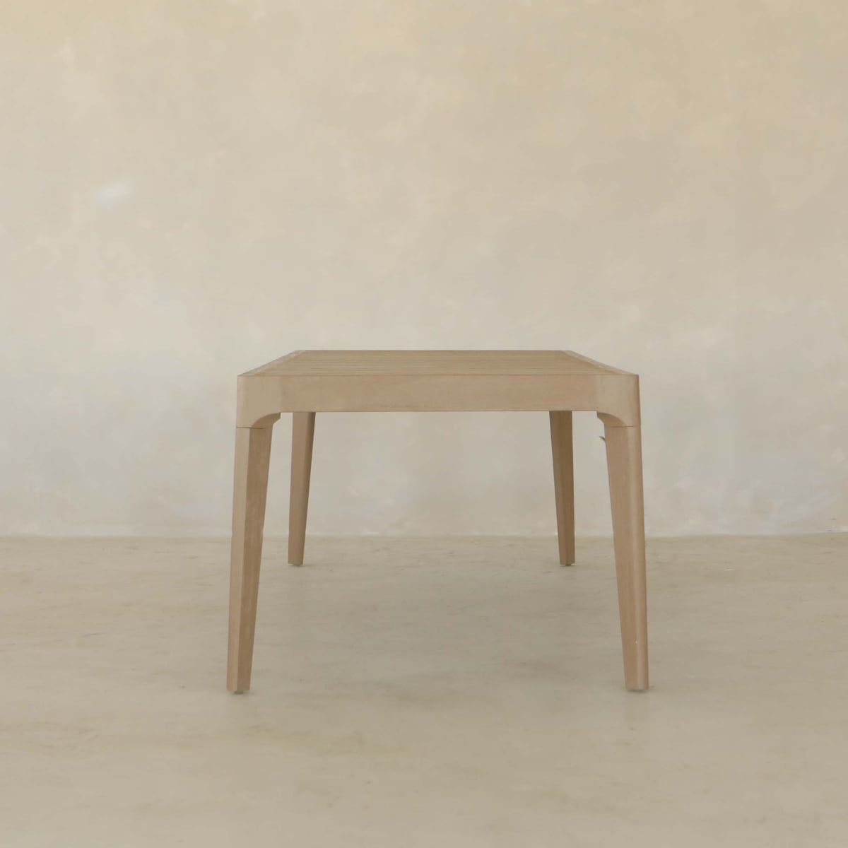 The Astier Outdoor Dining Table Set features a minimalist wooden design with four legs, inspired by French Modernist style, set against a plain, light-colored wall and floor background.