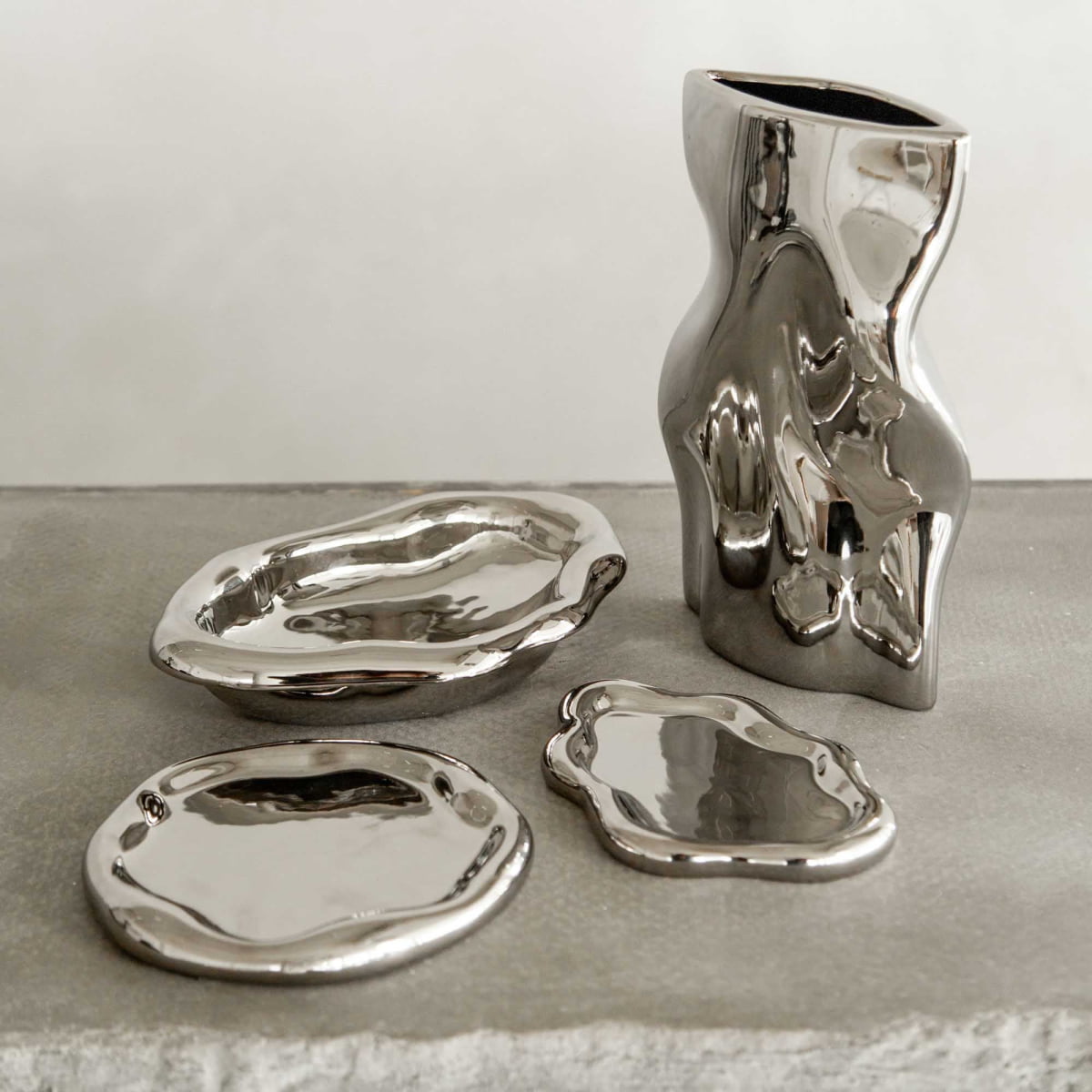 The Aura Dish, an irregularly shaped shiny silver plate, pairs with two other silver plates and a wavy abstract vase to create a contemporary display on a textured gray surface against a neutral backdrop.