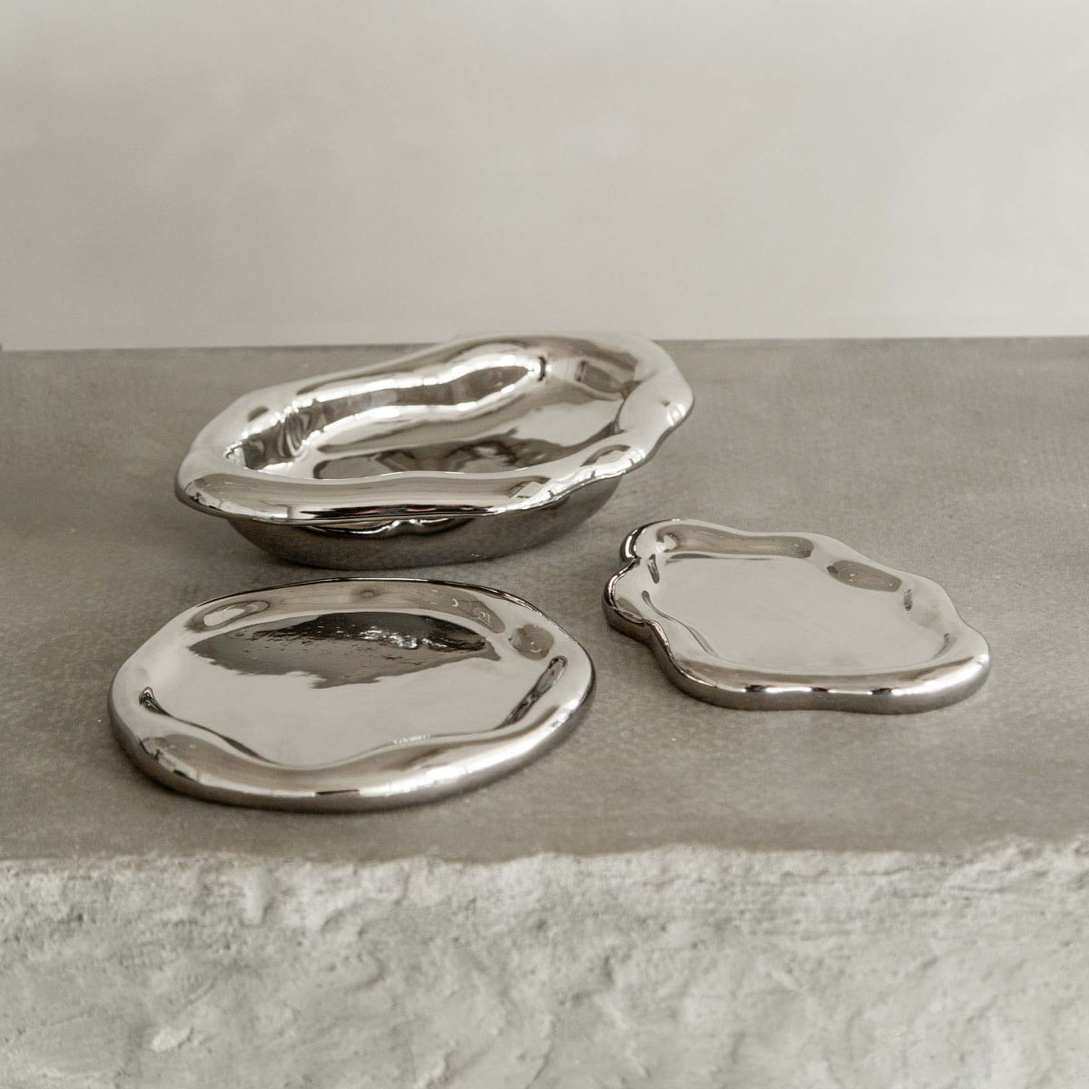 Three shiny silver trays from the Aura Dish collection, each with a unique sculptural shape and reflective finish, rest on textured stone against a neutral backdrop, capturing contemporary elegance.