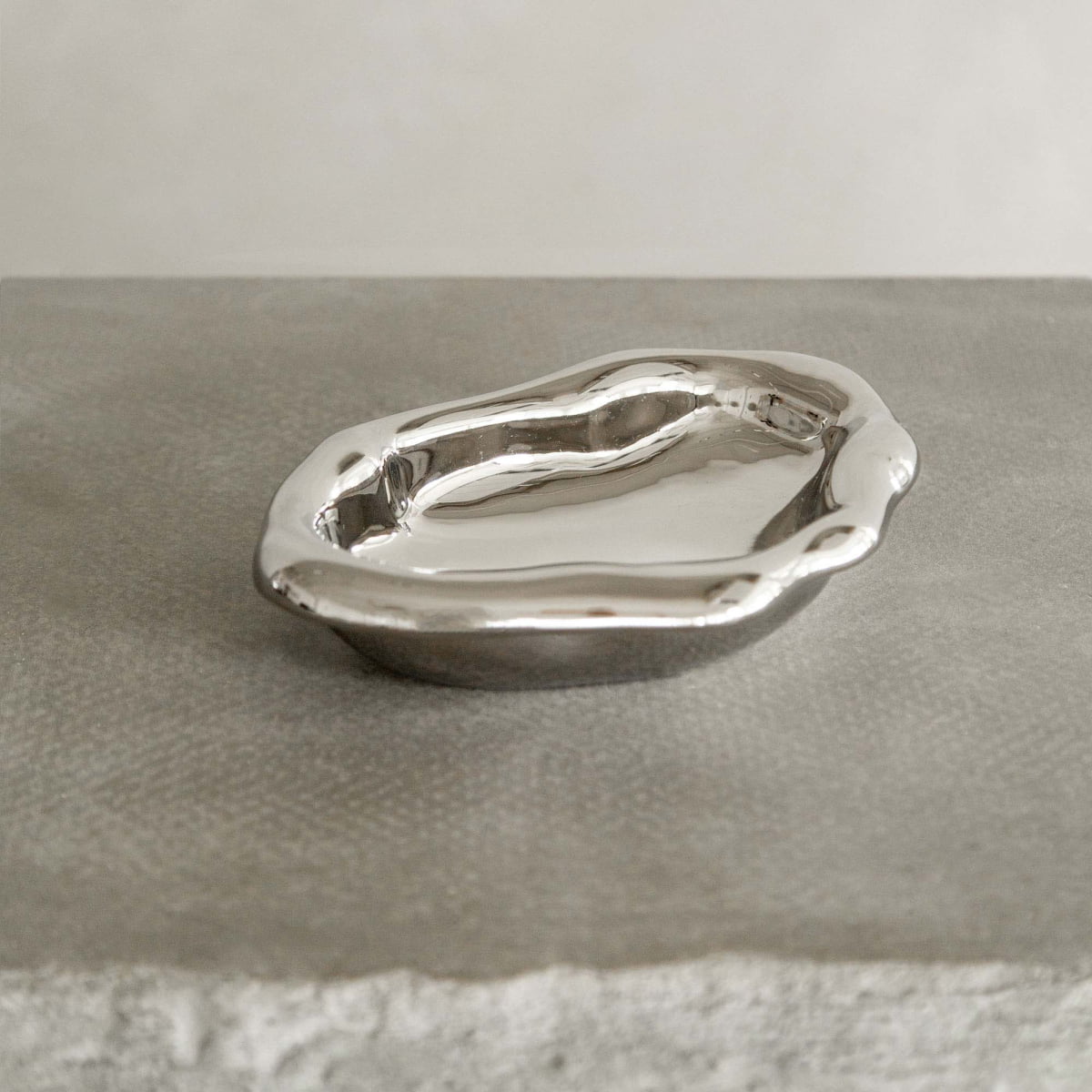 The Aura Dish, featuring a shiny finish and wavy edges, rests on a textured gray surface, highlighting its contemporary design against a neutral background.