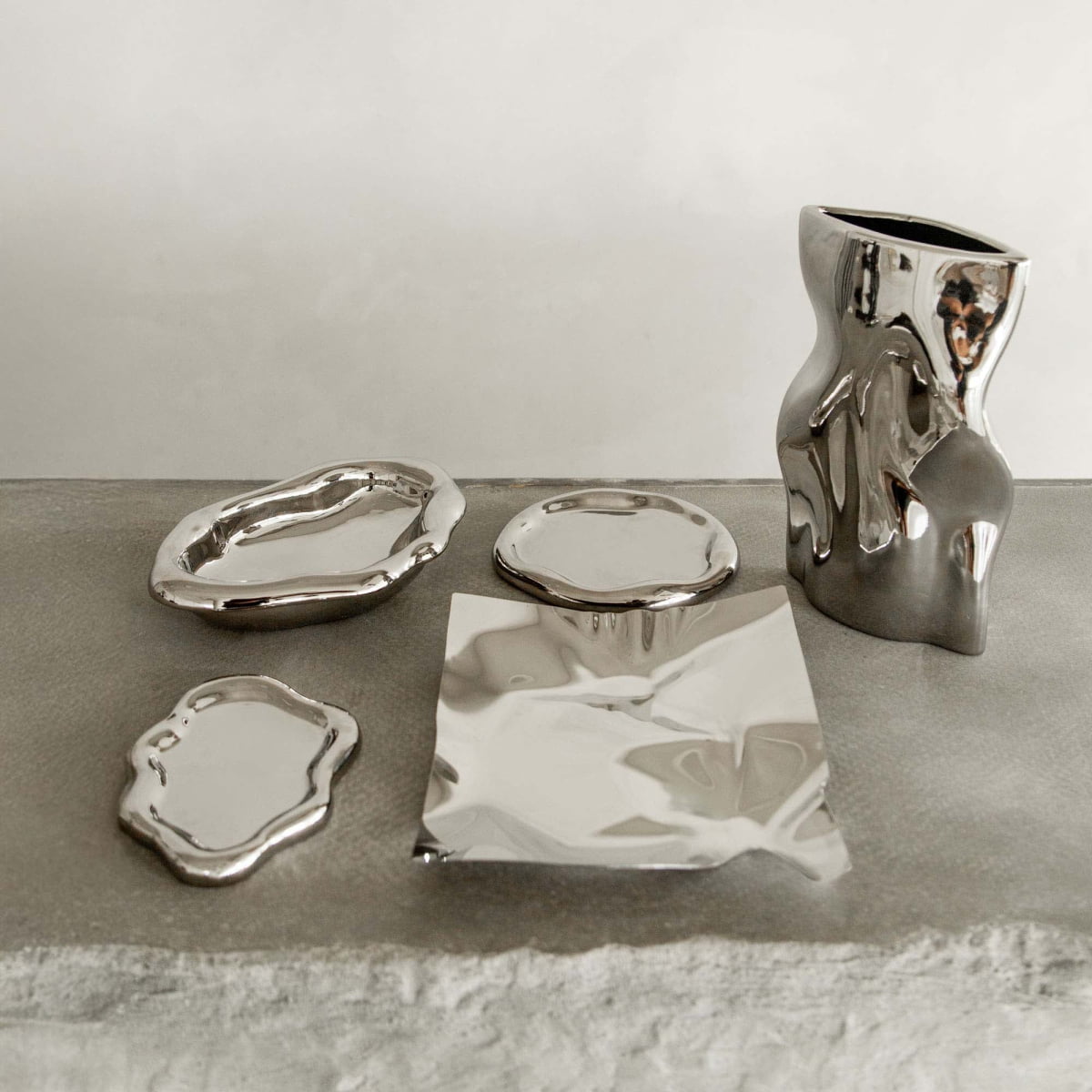 Five Aura Dish pieces with reflective silver surfaces and organic shapes are arranged on a textured gray surface, highlighting contemporary elegance against a plain, light background.