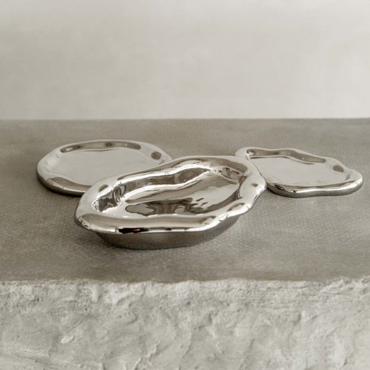 Three shiny silver Aura Dish trays, each with a unique shape, rest on a textured gray stone surface, showcasing contemporary elegance against a neutral background.