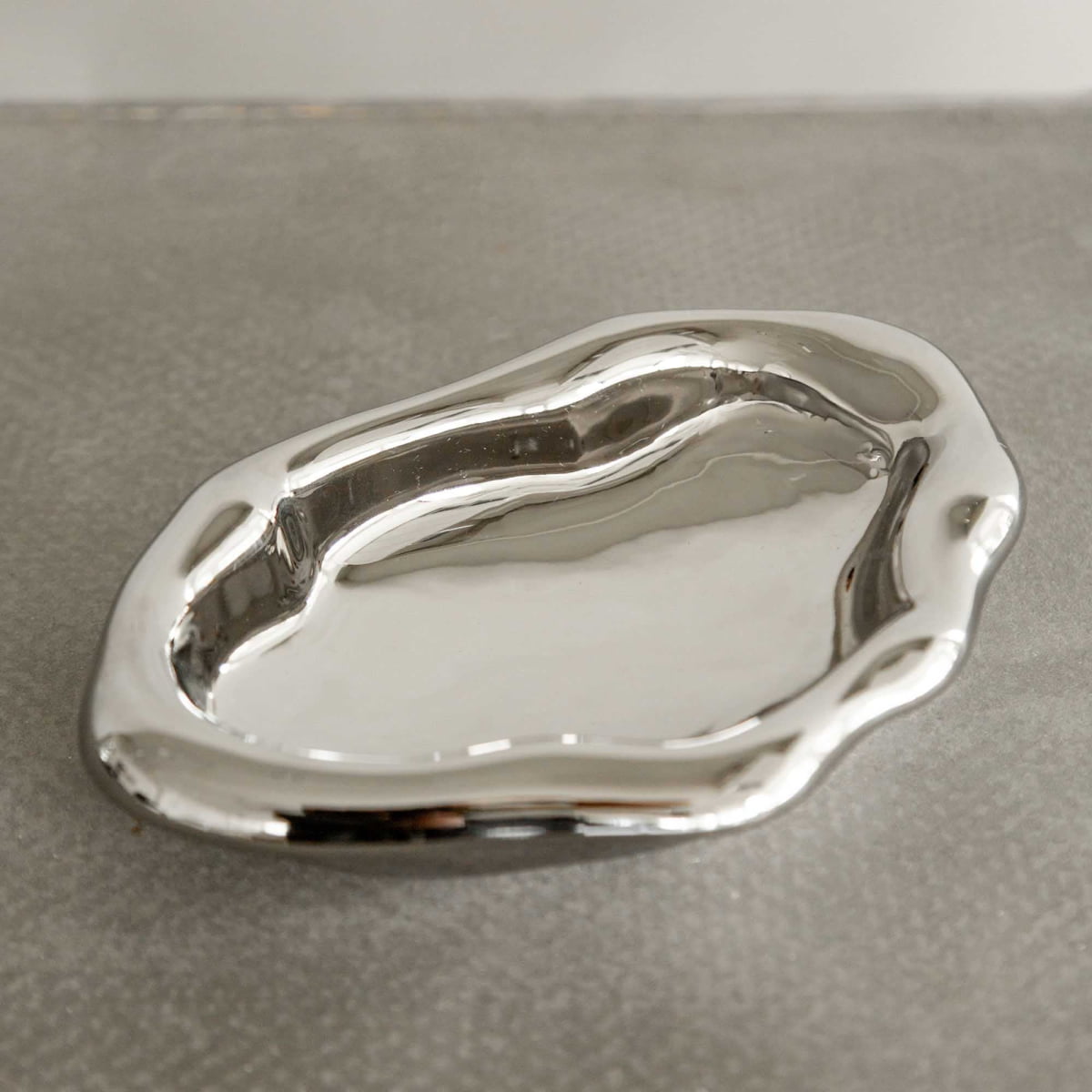 The Aura Dish features a shiny, silver finish and smooth, wavy edges. Its irregular sculptural shape adds contemporary elegance to any textured gray surface.