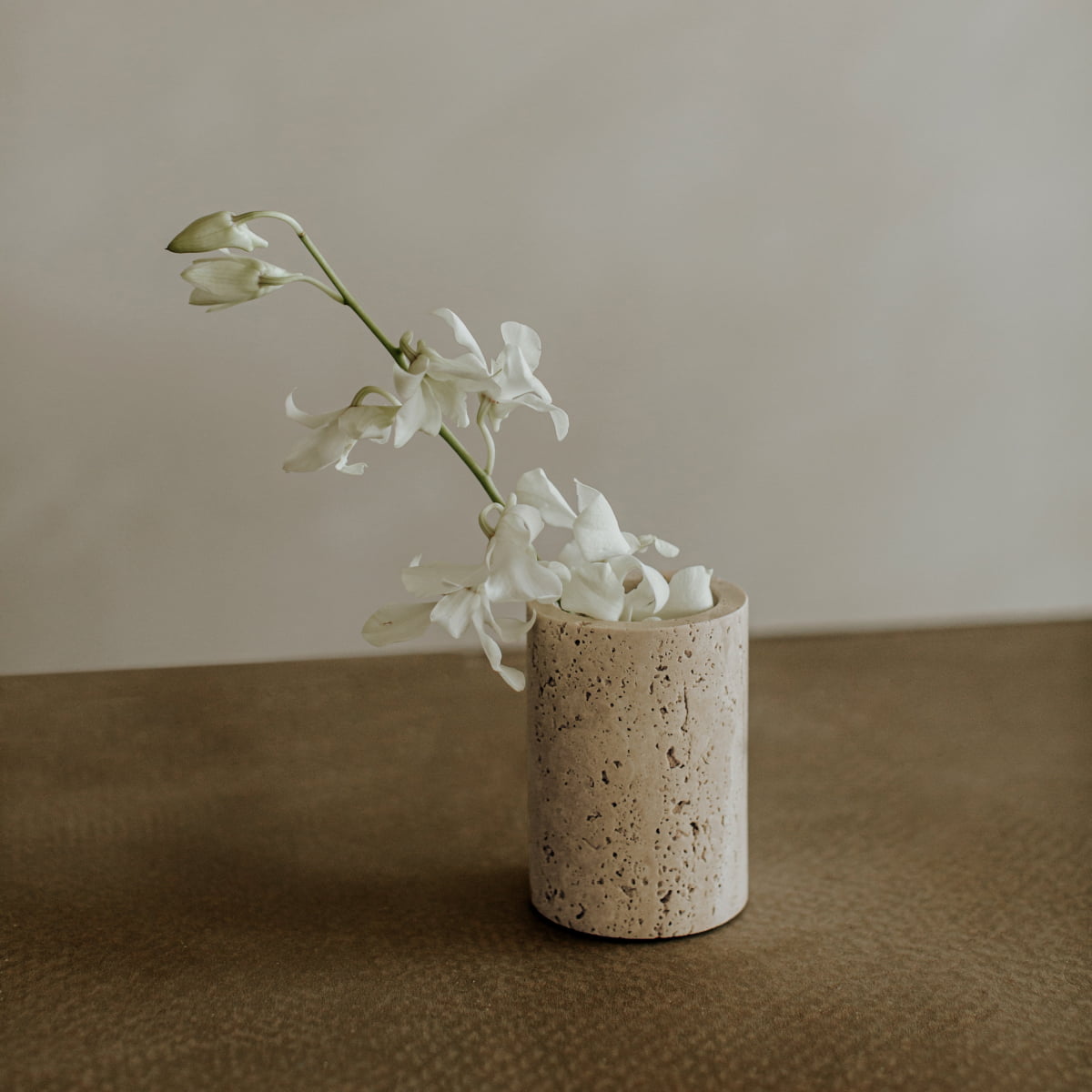 A single stem of white orchids in the Aurelia Goblet, a textured travertine vase, sits on a brown surface against a soft, neutral background.