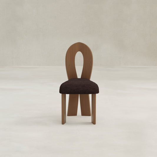 The Aveline Dining Chair features a unique teardrop backrest and a dark, textured upholstered seat, shown on a light floor with a neutral textured wall background.