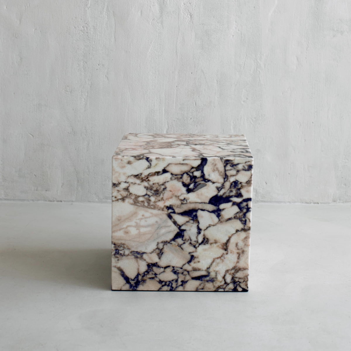 The Turkish Calacatta Plinth Cube, with beige, cream, and black veining, rests on a smooth light gray floor beside a textured light wall.