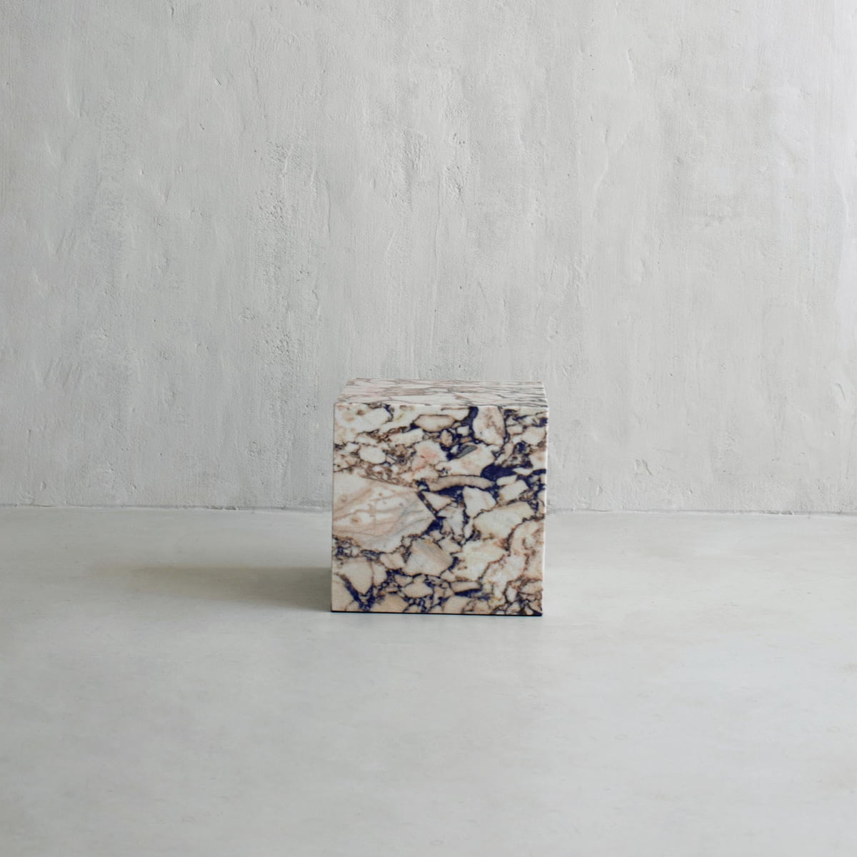 The Turkish Calacatta Plinth Cube, with a beige and dark veined marble pattern, sits on a smooth light gray floor against a plain textured wall.