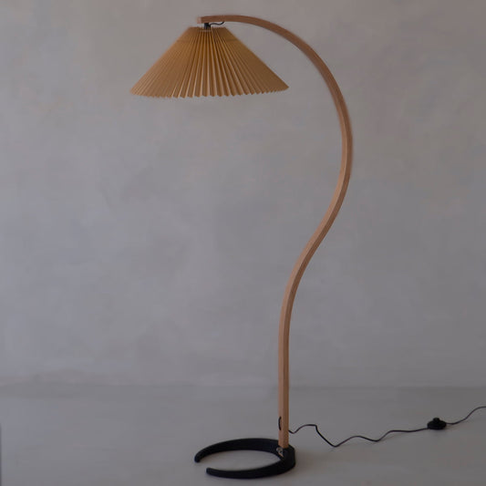 The Bailey Lamp features a curved wooden stand, pleated brown shade, and black semicircular base, bringing modern style to any light-colored floor and neutral wall. A black cord extends from the base for a sleek finishing touch.