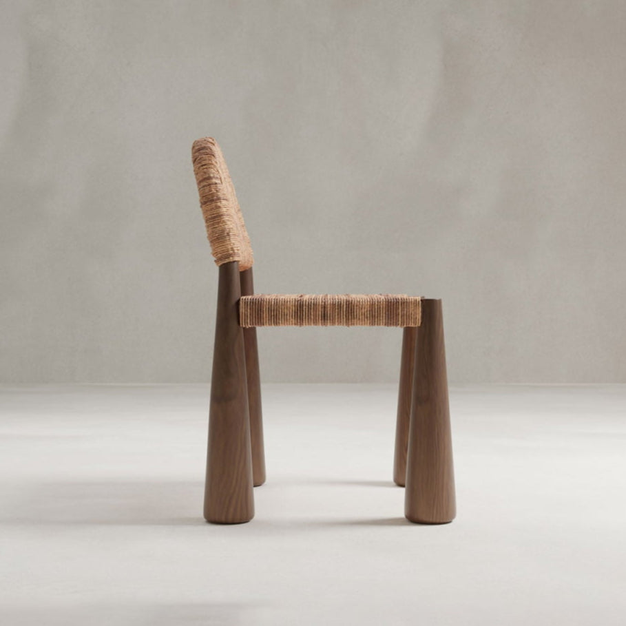 The Bardo Chair features a modern handcrafted design with a woven seat and backrest, wooden frame, and cylindrical legs, highlighting artisanal craftsmanship in side profile against a plain light gray background.