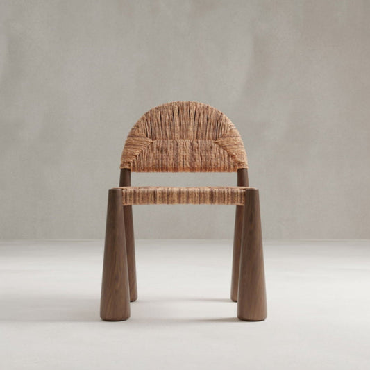 The Bardo Chair features artisanal craftsmanship with a rounded woven seat and backrest, supported by three sturdy brown wooden legs, all set against a plain light gray background.