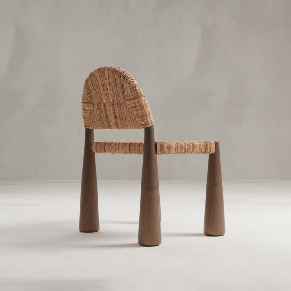 The Bardo Chair features a contemporary, handcrafted design with a woven natural-fiber backrest and seat, plus thick rounded wooden legs, shown against a plain light gray background.
