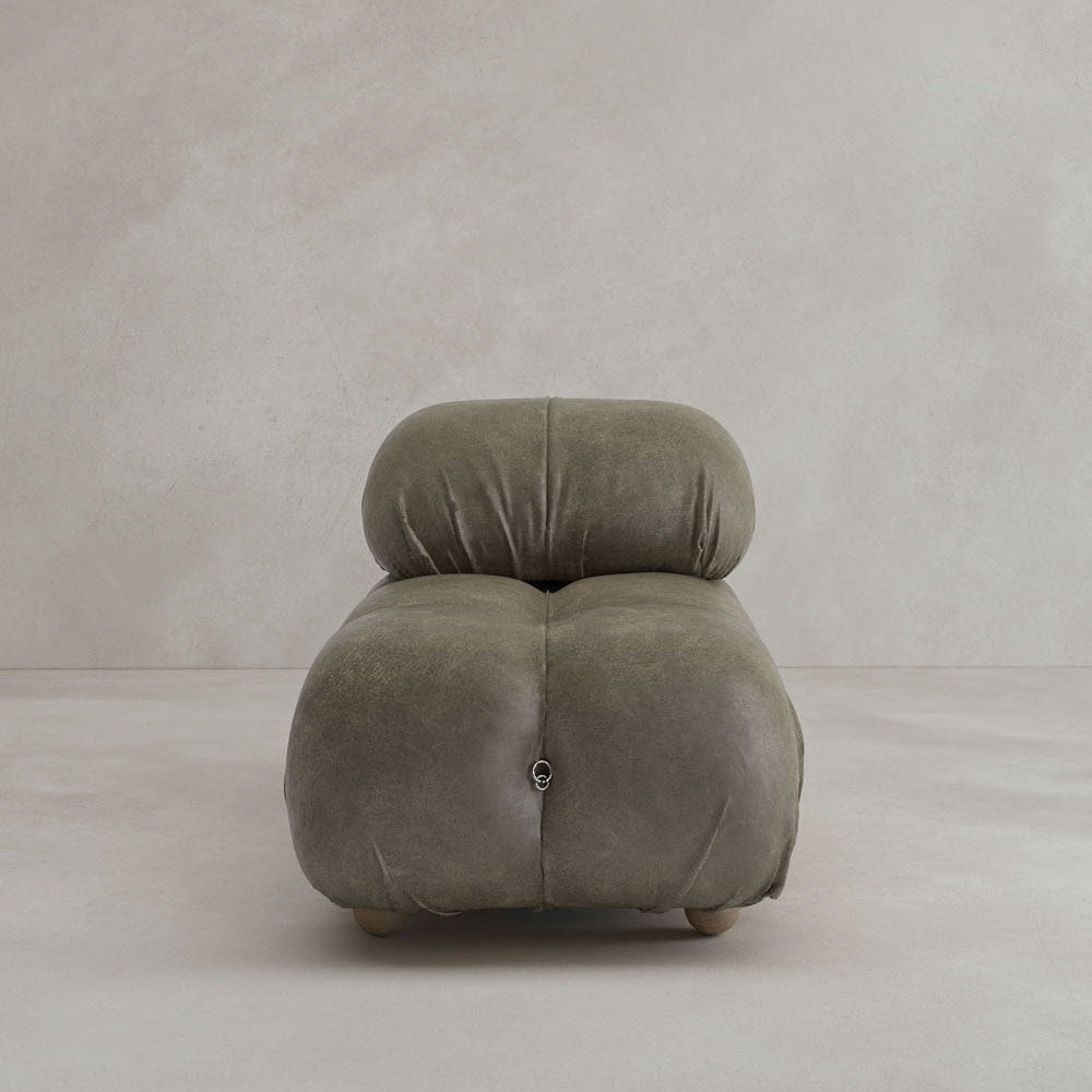 A plush, olive-green Belmont Chair 2/3Rd Modular Sectional with rounded edges and tufted upholstery sits on a light, neutral floor against a textured wall—an iconic classic for any space.