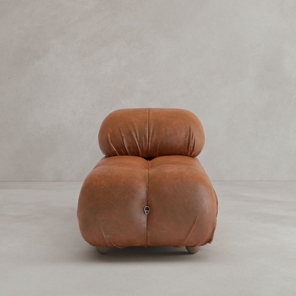 The Belmont Chair 2/3Rd Modular Sectional, featuring marshmallow-like curves and tufted brown leather upholstery, sits against a plain light gray wall and floor.