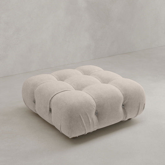 The Belmont Ottoman, a square light beige tufted piece with a plush cushioned surface and modular design, sits on a smooth light gray floor against a plain light gray wall.