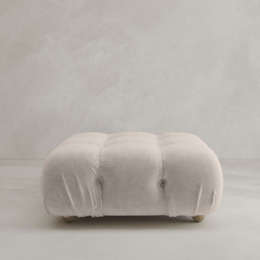 The Belmont Ottoman, a square, tufted piece upholstered in light beige fabric with refined button detailing and classic style, is set against a plain light gray background.