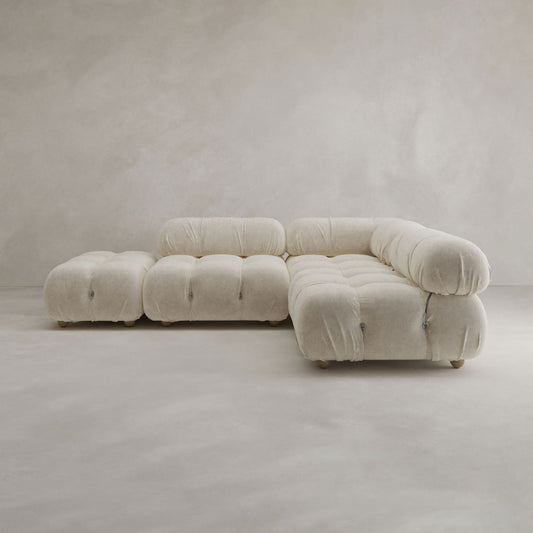 The Belmont Sofa Combination B features modular L-shaped seating, an ottoman, and plush, tufted cream fabric. Its rounded edges and soft upholstery create a modern look in any minimal, neutral-toned room.