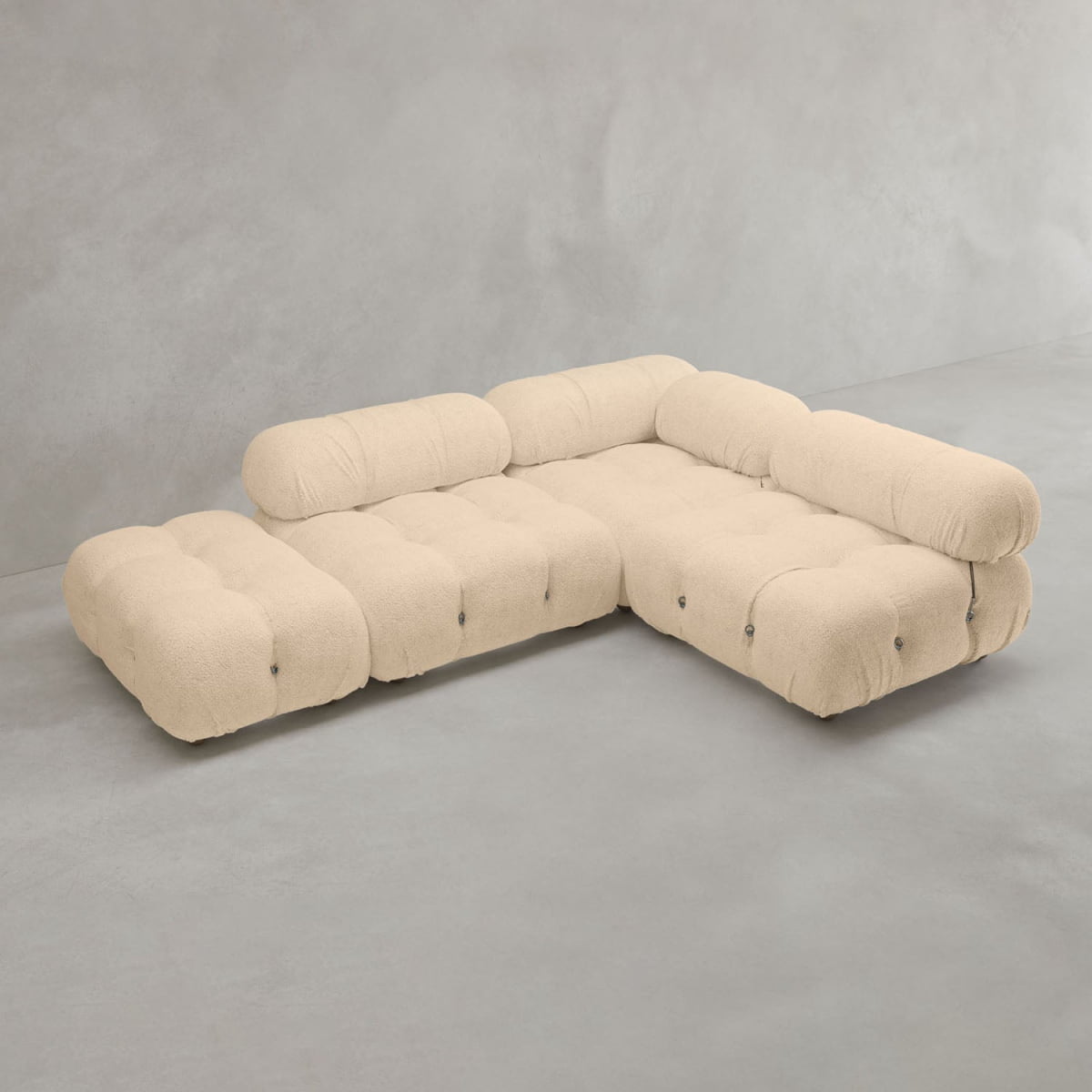 The Belmont Sofa Combination B, an L-shaped modular design with plush tufted cream cushions and rounded edges, sits on a smooth light gray floor against a matching gray wall.