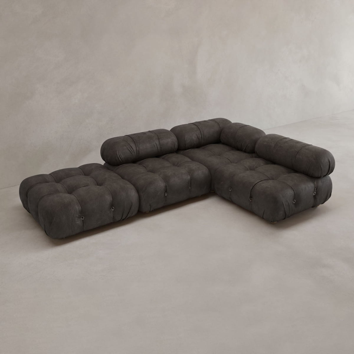 The Belmont Sofa Combination C, a low-profile dark gray modular sectional with tufted cushions and soft curves, is arranged on a light concrete floor against a plain neutral wall.