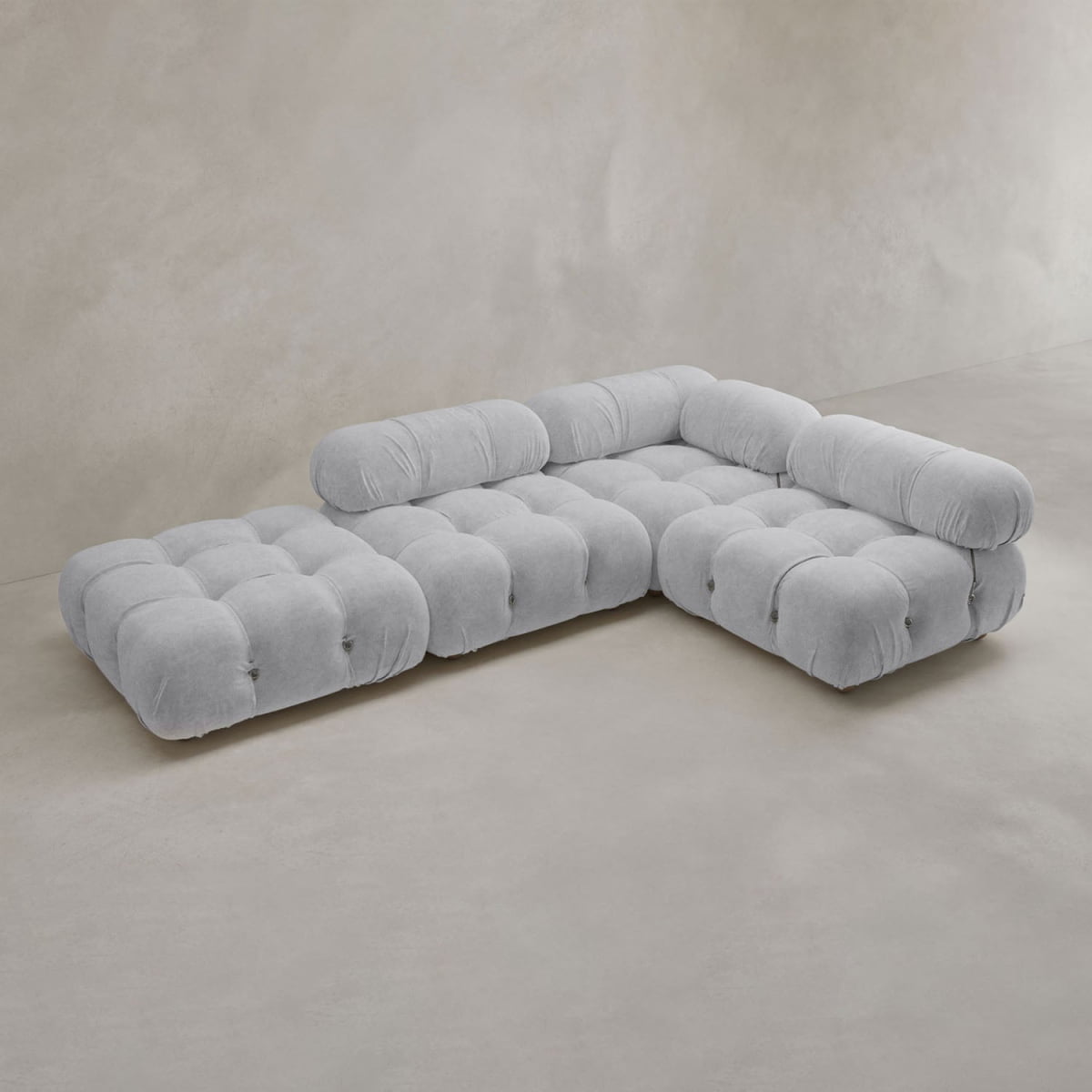 The Belmont Sofa Combination C is a modern, light gray tufted sectional with classic appeal, rounded armrests, and marshmallow-like curves, set on a smooth neutral-toned floor against a plain beige wall.