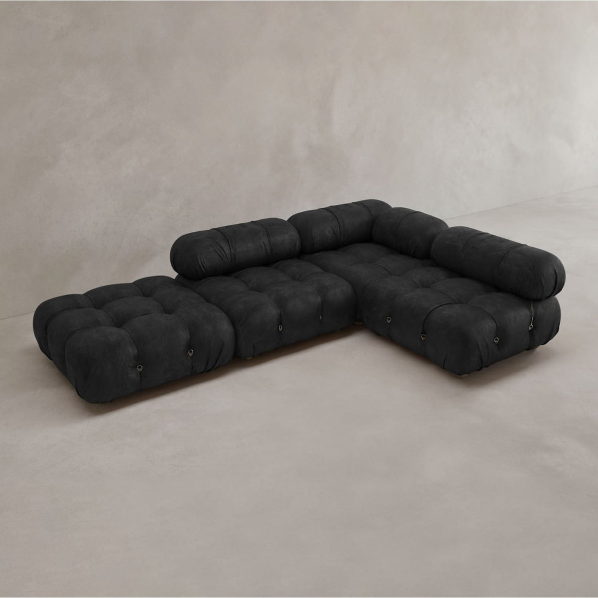 The Belmont Sofa Combination C is a modern, modular black sectional with tufted cushions, marshmallow-like curves, and cylindrical bolsters, arranged in an L-shape against a plain light-colored wall and floor.
