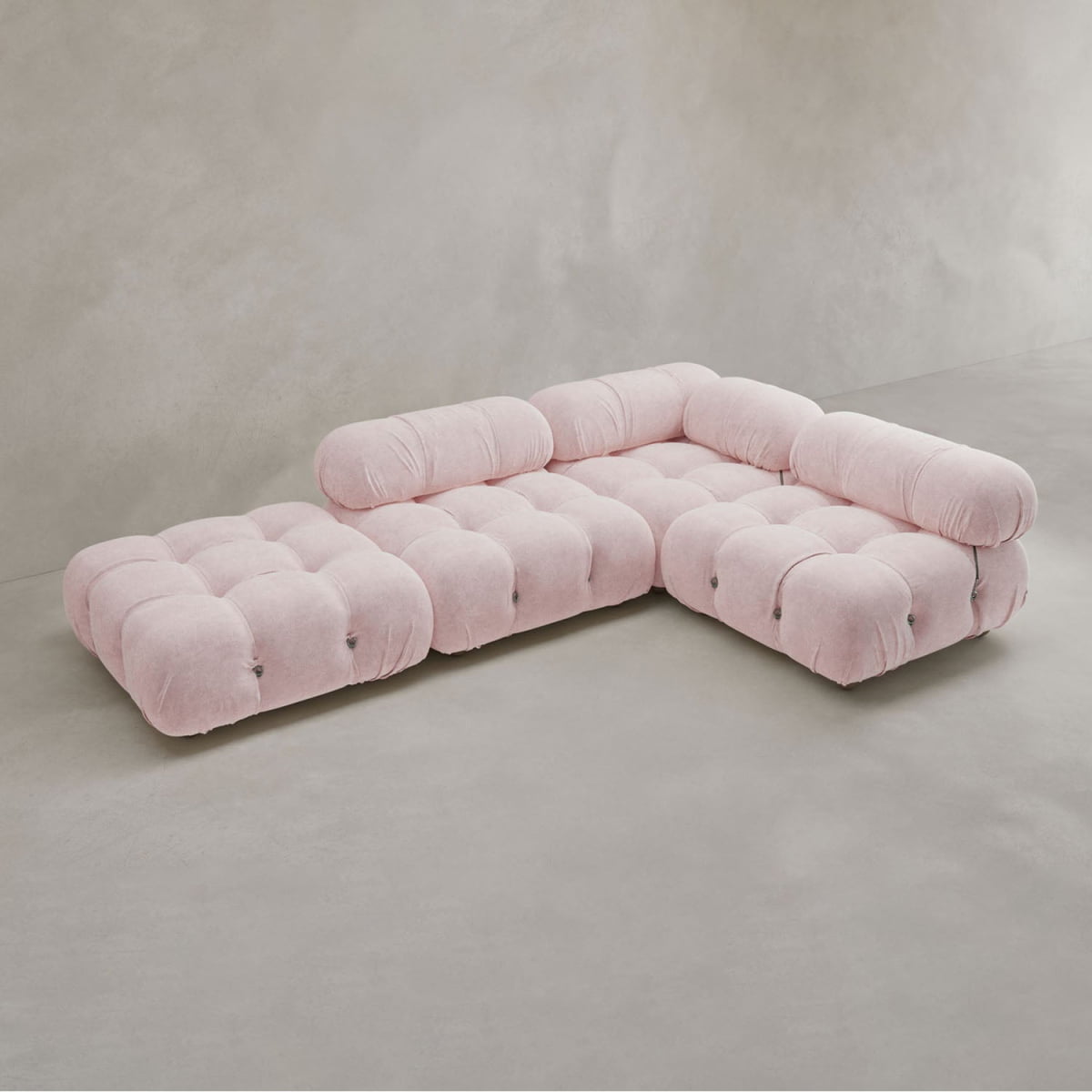 The Belmont Sofa Combination C is a modular, L-shaped pink sofa with tufted cushions and marshmallow-like curves, positioned on a smooth light grey floor against a plain neutral wall.