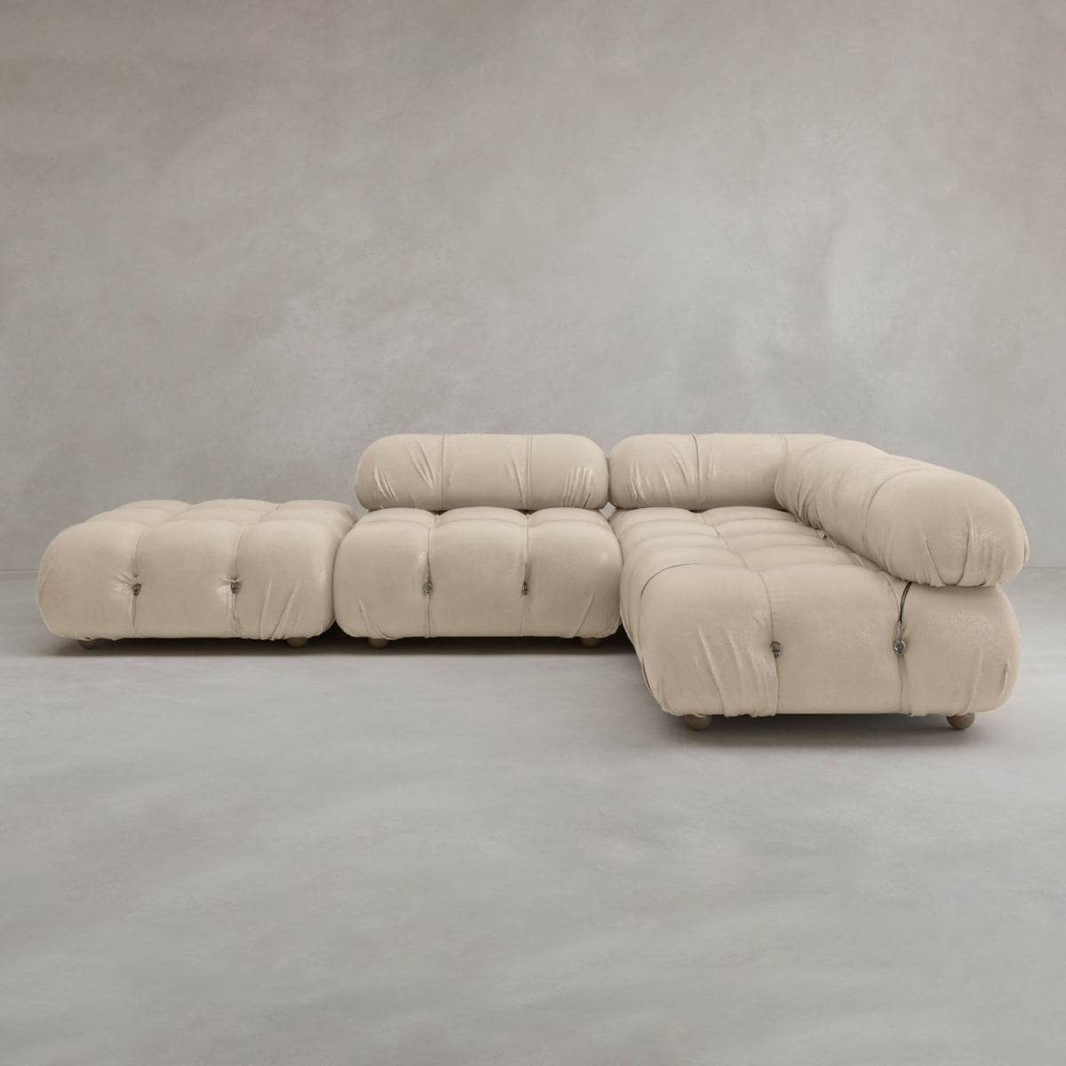 The Belmont Sofa Combination C, a modern, modular beige sectional with tufted cushions and rounded edges, features marshmallow-like curves and sits on a light gray floor against a neutral gray wall.