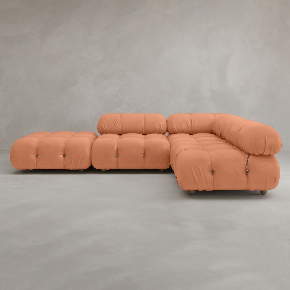 The Belmont Sofa Combination C is a modular orange sofa with classic tufted, marshmallow-like curves and an L-shaped design, set on a smooth light gray floor against a plain gray wall.