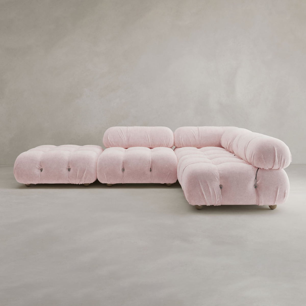The Belmont Sofa Combination C, a modern modular sectional in light pink with marshmallow-like curves and tufted cushions, features a chaise lounge and matching ottoman, positioned against a plain textured grey background.