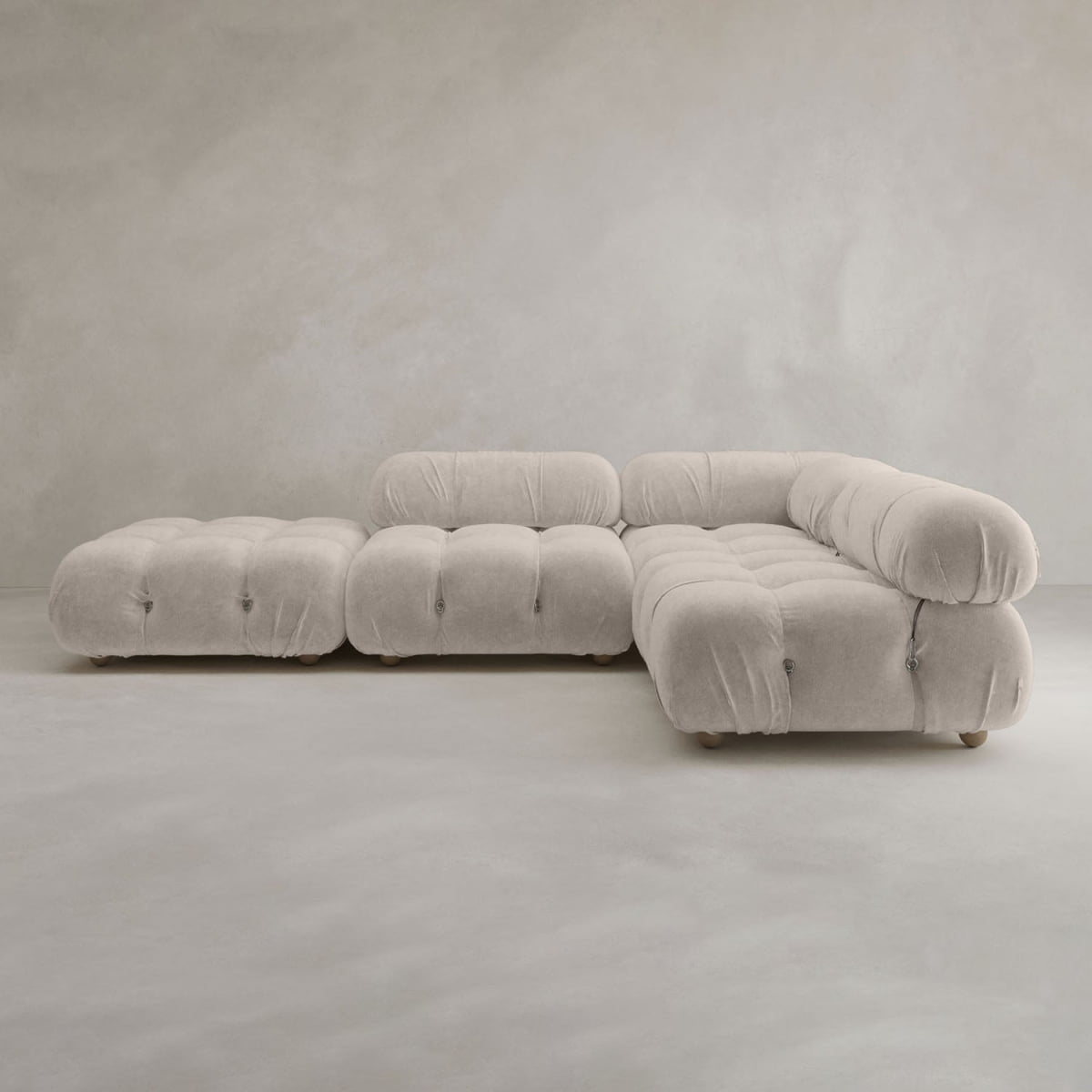 The Belmont Sofa Combination C is a modern, light beige sectional with tufted, marshmallow-like curves and an ottoman, set against a neutral wall and floor.