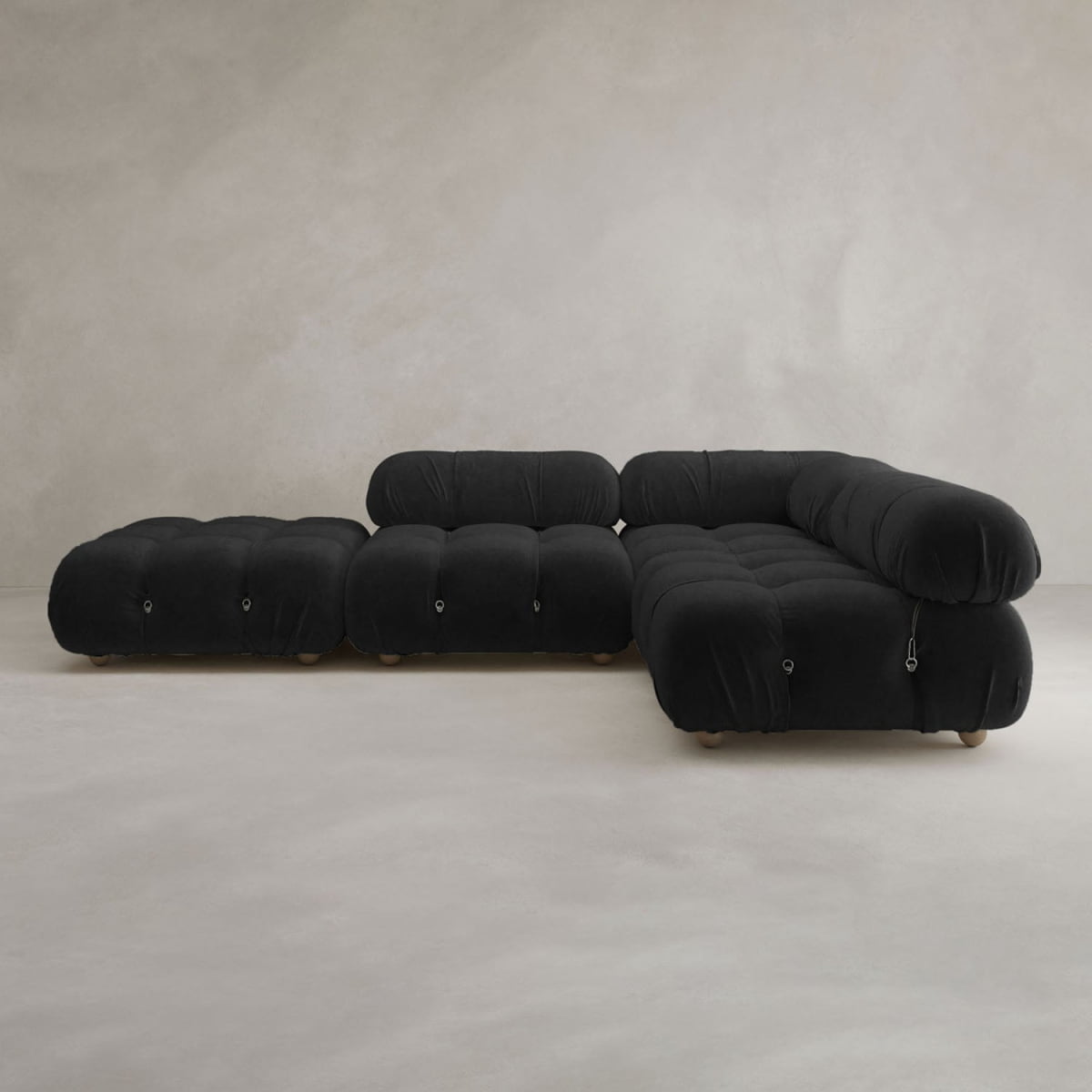 The Belmont Sofa Combination C, a modern black sectional with curved, tufted cushions, sits next to a matching ottoman against a textured neutral wall and light floor.