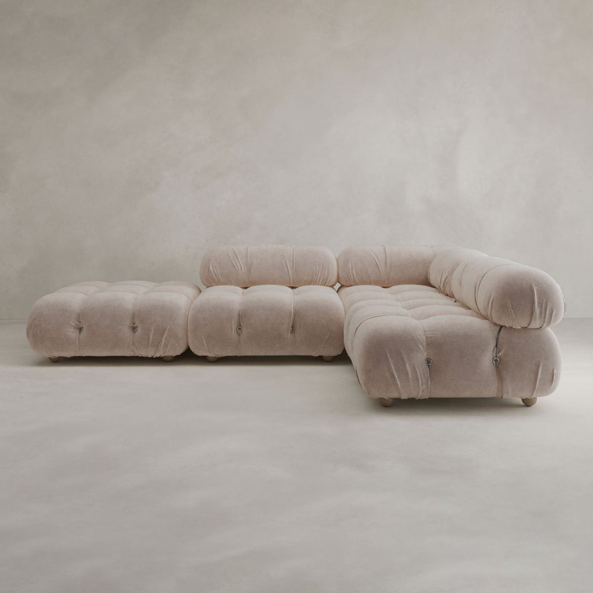 The Belmont Sofa Combination C, a modern modular beige sectional with plush tufted cushions and rounded curves, sits in a minimalist room featuring light, textured walls and a matching floor.