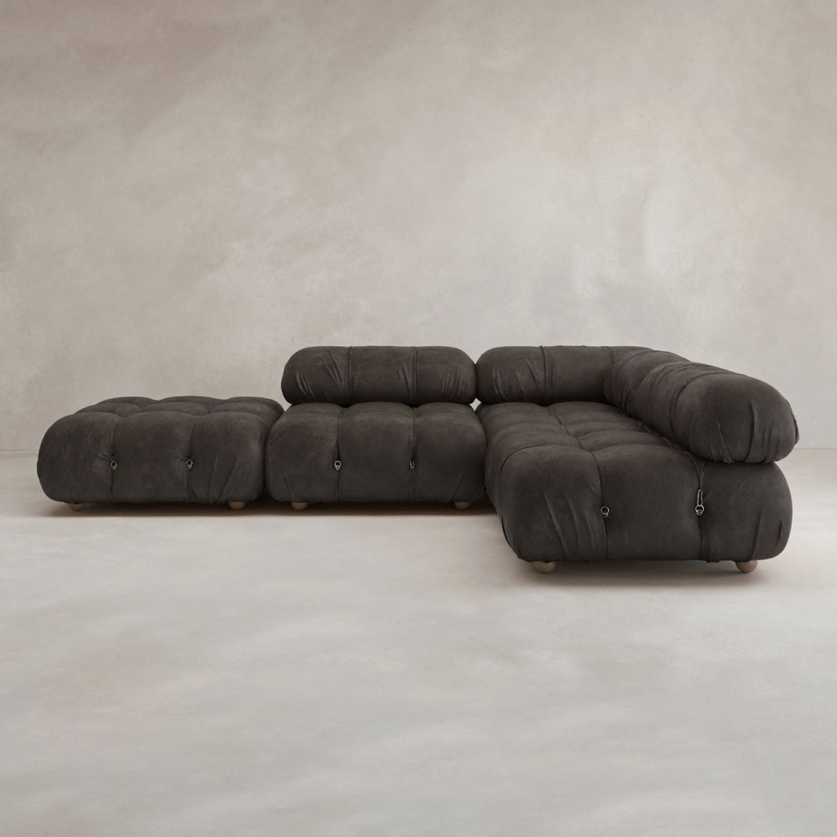 The Belmont Sofa Combination C is a modular, dark gray tufted sectional with marshmallow-like curves and an ottoman, set against a plain light-colored wall and floor.