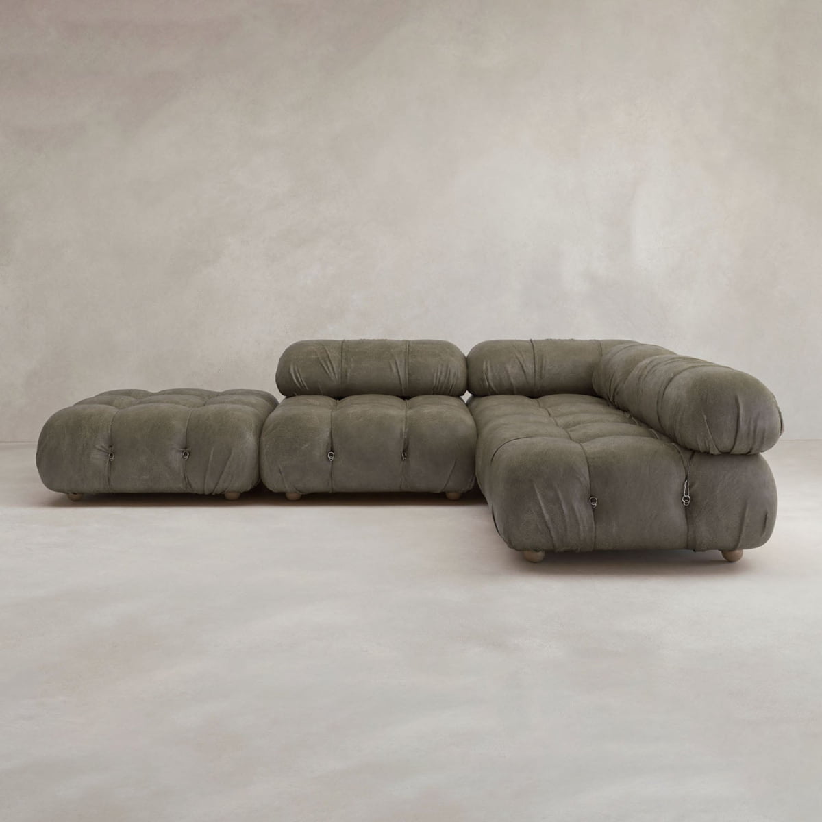 The Belmont Sofa Combination C is a modern olive-green sectional with marshmallow-like curves, tufted cushions, and rounded edges. Its modular design features a chaise lounge and matching ottoman, set against a neutral textured wall and light floor.