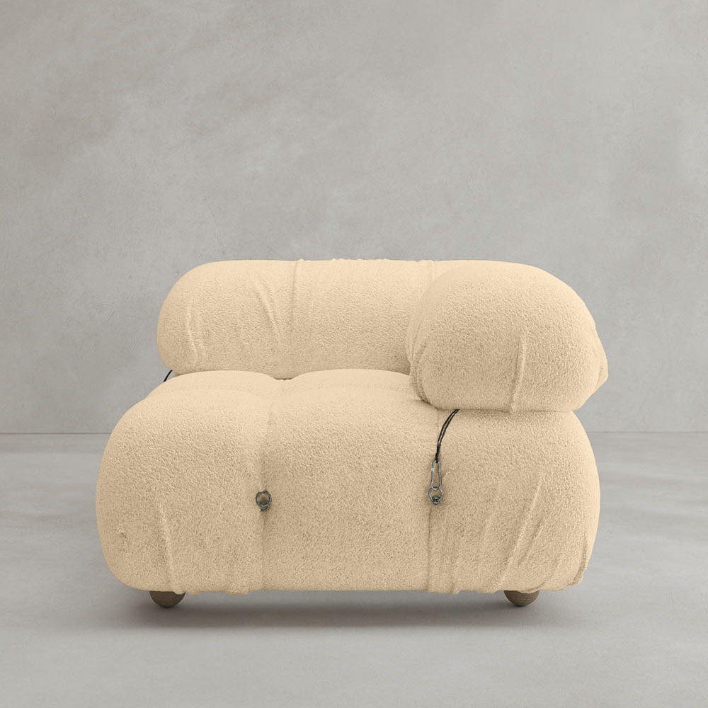 The Belmont Sofa Left Armrest features a modern modular design with soft, textured beige fabric, rounded edges, visible tufting, and exposed metal accents, shown against a plain gray background.