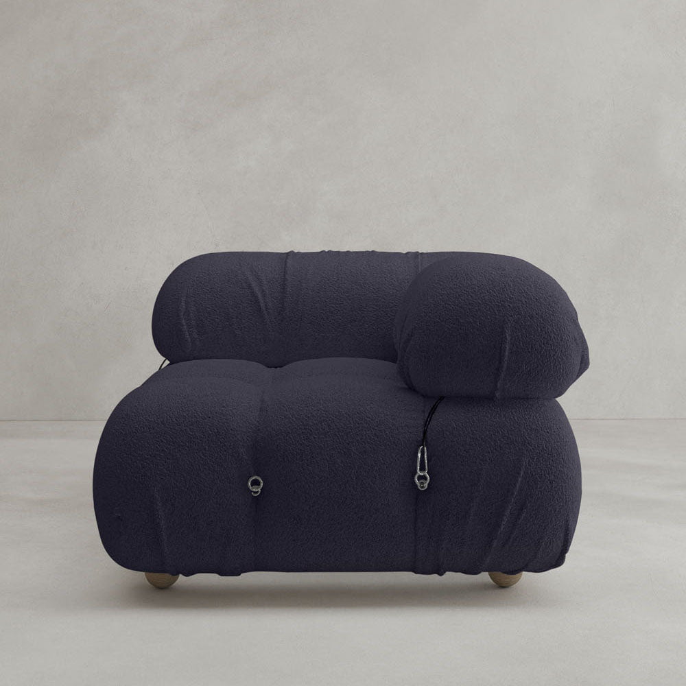 The Belmont Sofa Left Armrest features a modern, deep navy look with plush, rounded design and soft, textured fabric. Inspired by classic styles, it has metal ring and zipper accents on the sides against a plain light gray background.