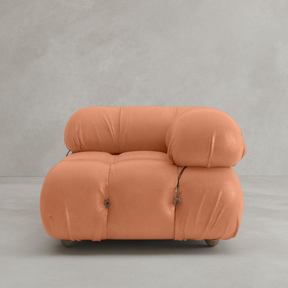 The Belmont Sofa Left Armrest, featuring plush marshmallow-like curves and soft orange upholstery with visible button tufts and metal fasteners, sits against a plain light gray wall and floor background.