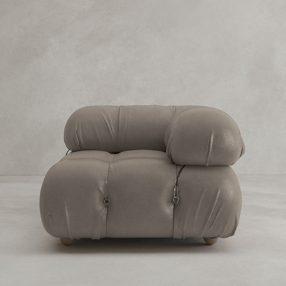 The Belmont Sofa Left Armrest in plush taupe, featuring a rounded, tufted design and visible seams, offers an iconic classic look against a plain light gray wall and floor.