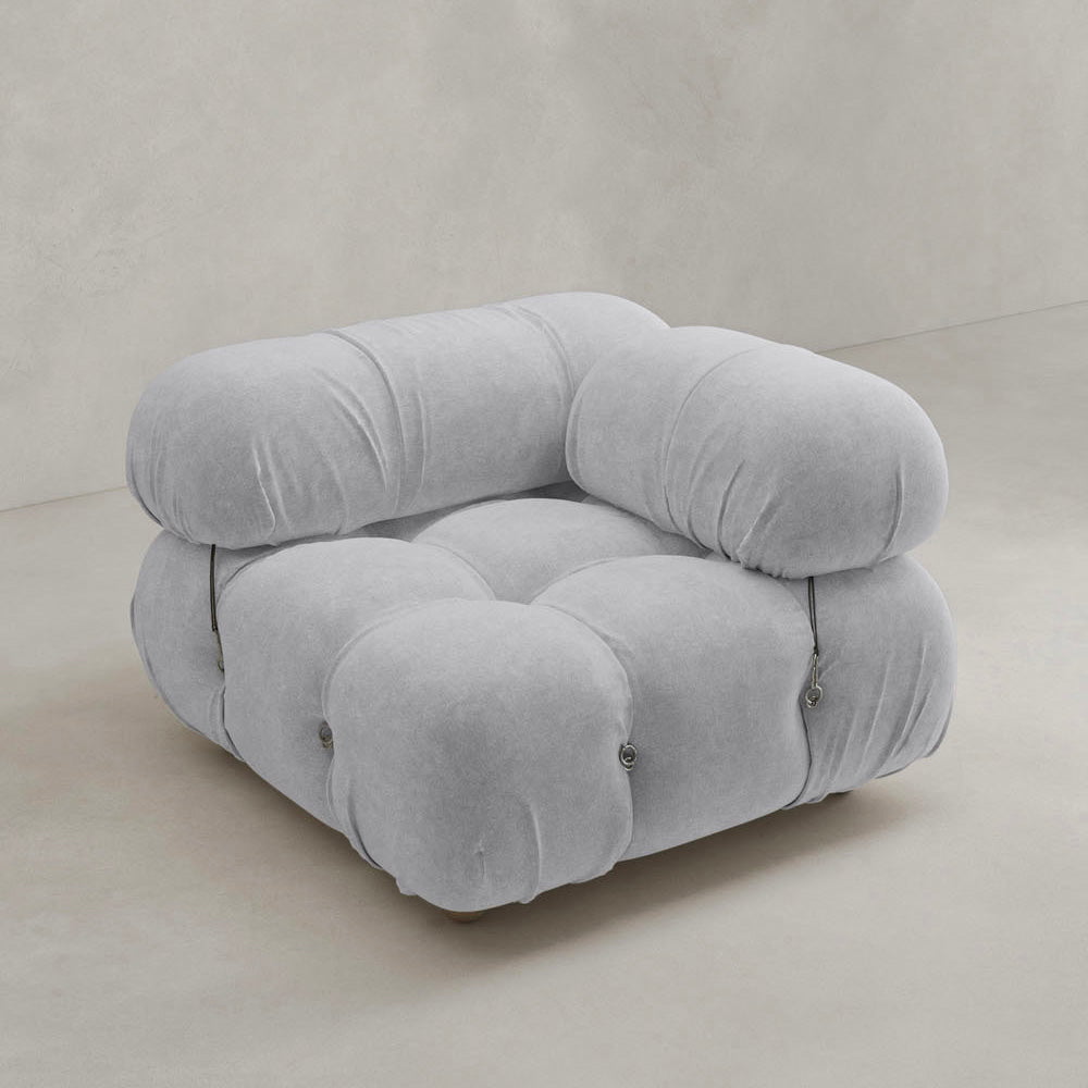 The Belmont Sofa Left Armrest features plush, tufted light gray upholstery with soft, rounded edges and marshmallow-like curves, set on a neutral-toned floor against a plain beige wall.