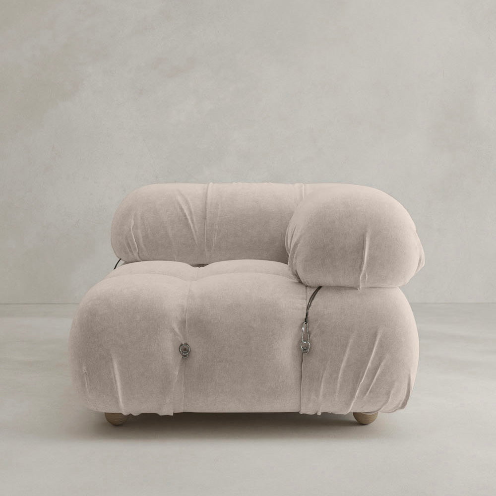 The Belmont Sofa Left Armrest features plush, light beige upholstery with marshmallow-like curves, subtle button tufting, and small wooden legs, perfect against a neutral textured wall and floor.