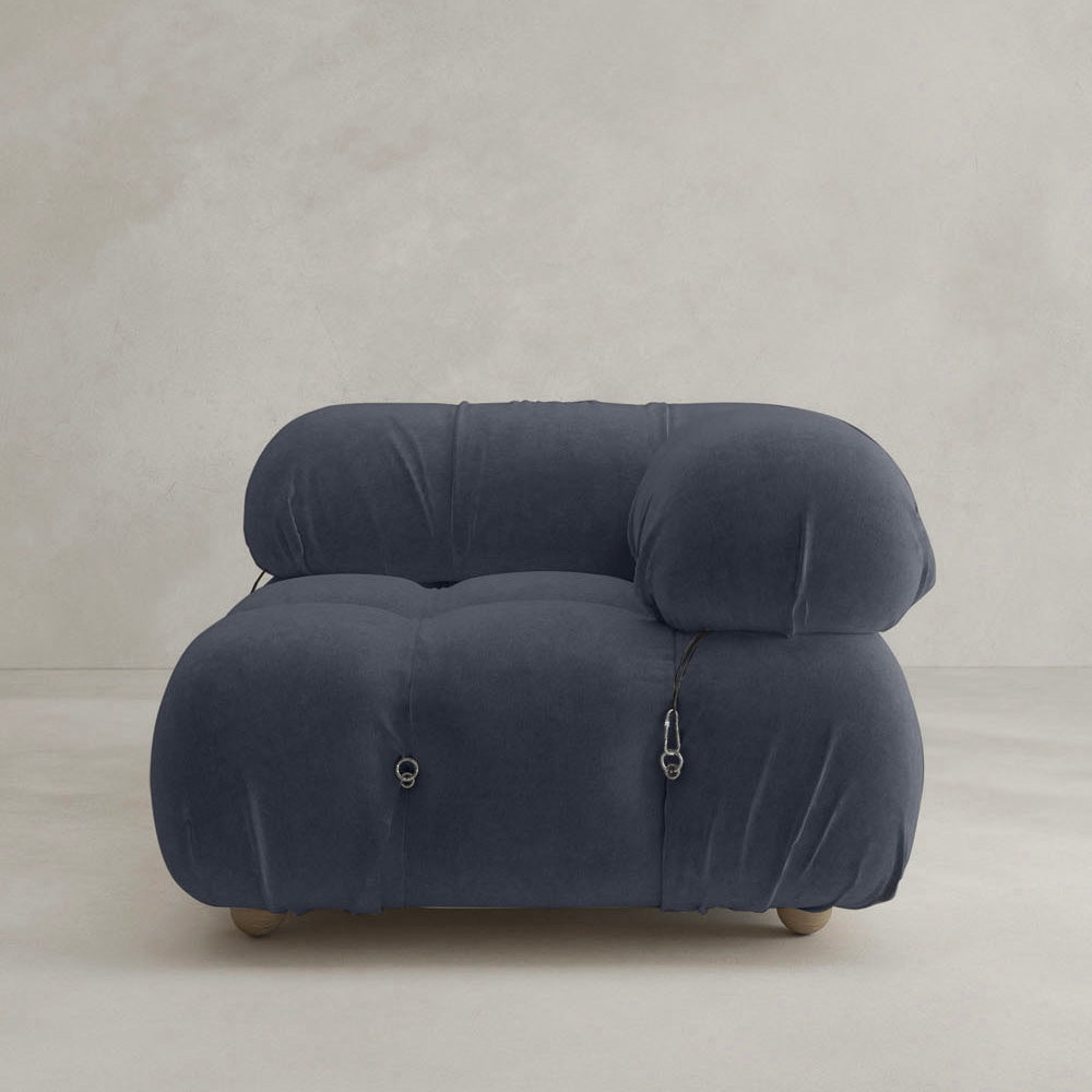 The Belmont Sofa Left Armrest features dark blue upholstery, rounded cushions, and visible metal fasteners, highlighting classic style in a minimalist room with light-colored walls and flooring.