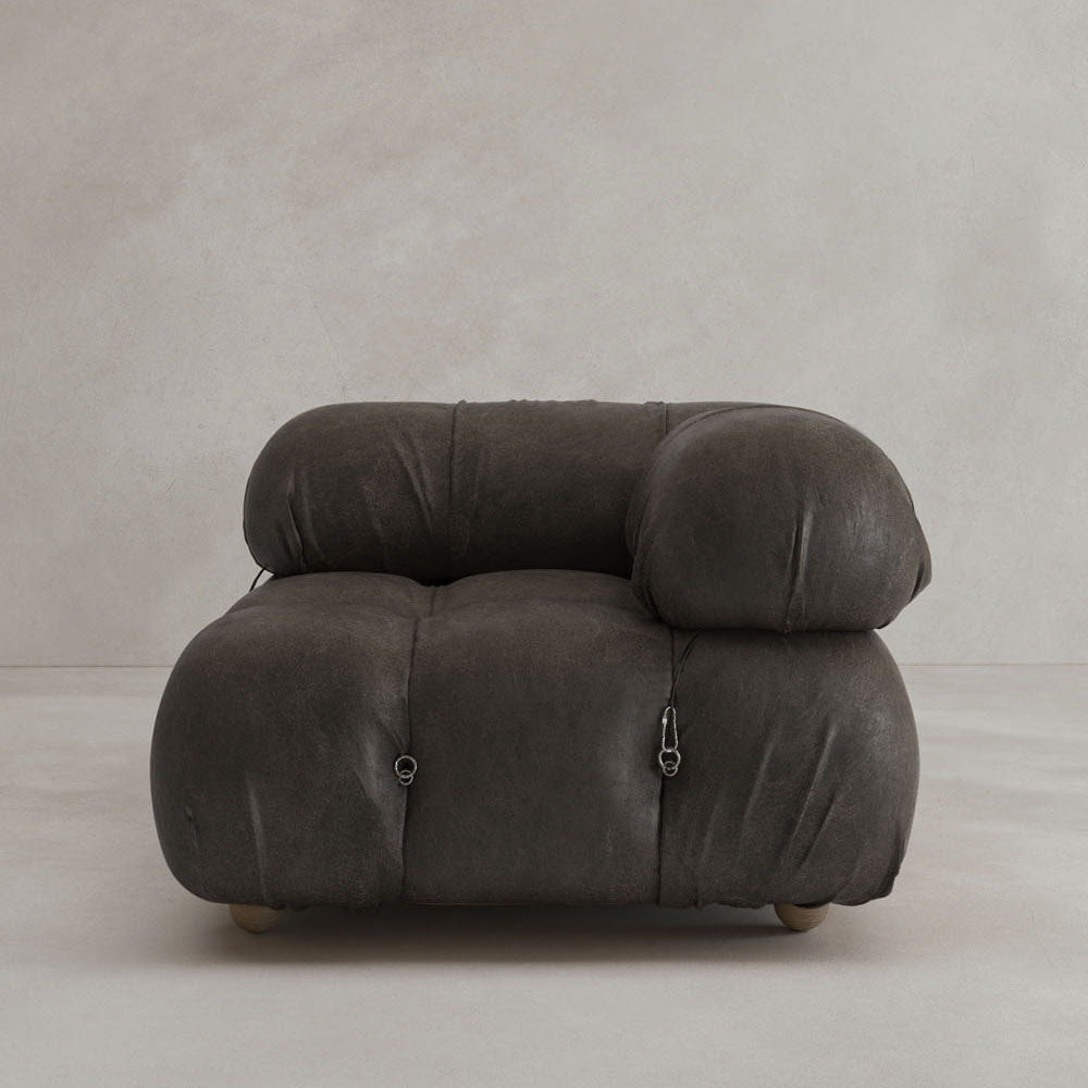 The Belmont Sofa Left Armrest features a modern, oversized design in dark gray with rounded, tufted cushions and visible metal fasteners, highlighting its modular style against a plain, light wall and floor.