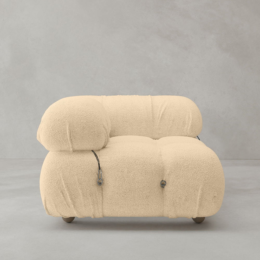 The Belmont Sofa Right Armrest is a modern beige plush seat with a rounded, overstuffed modular design and visible black straps, set against a plain light gray wall and floor.