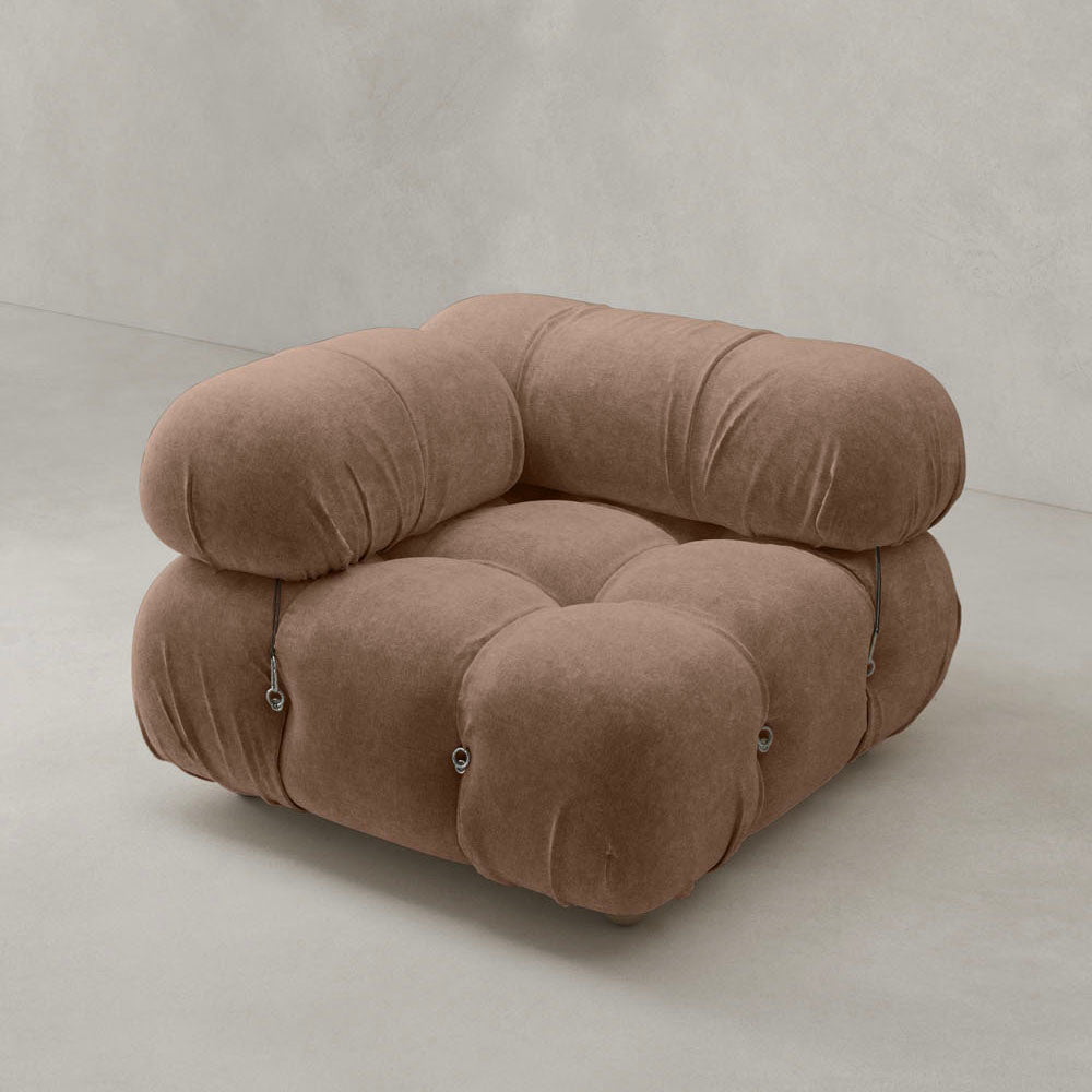 The Belmont Sofa Right Armrest, featuring a plush pillow-like design and tufted brown upholstery with rounded edges, brings iconic style to any space, shown here on a light gray floor against a plain, light-toned wall.