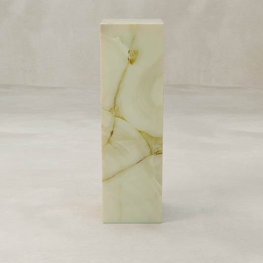 The Blanchaire Onyx Tall Tower Plinth is a tall, rectangular pedestal crafted from light beige marble with brown and gold veining, featuring a luminous finish that makes it a striking accent atop a smooth, light gray surface against a neutral background.