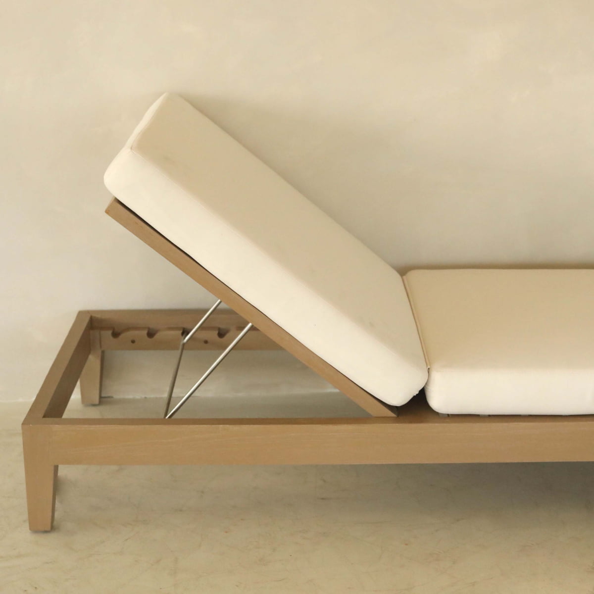 The Bourdain Patio Chaise Lounge with white cushions features an ergonomic, adjustable design and stands upright against a plain beige wall, providing stylish and comfortable outdoor seating.