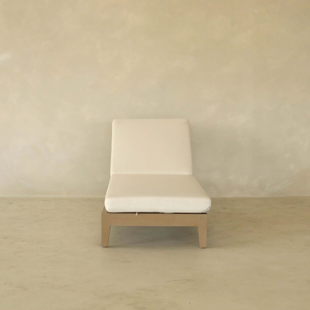 The Bourdain Patio Chaise Lounge features a minimalist, armless design with a white cushion and light wooden legs, providing outdoor comfort on a neutral floor by a beige textured wall.
