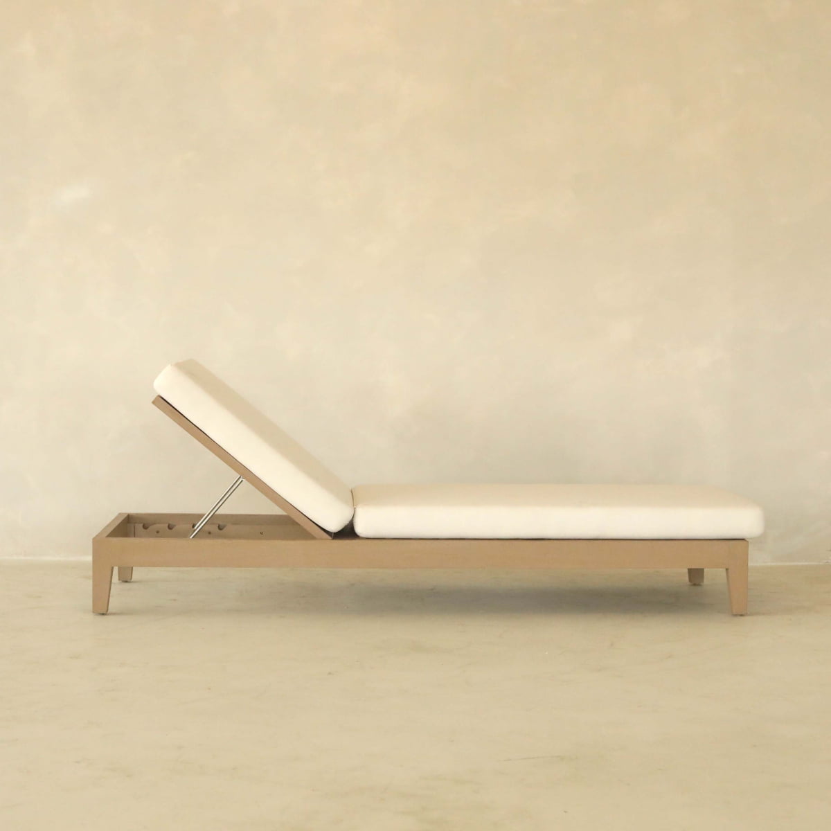 The Bourdain Patio Chaise Lounge features a minimalist beige design with a simple wooden frame and adjustable backrest, providing outdoor comfort. Its shown against a plain, light-colored wall and floor.